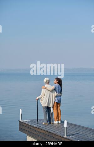 Vista posteriore della nonna e della nipote che si abbracciano mentre si trovano sul molo e si divertono a passeggiare insieme sul lungomare Foto Stock