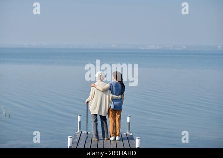 Vista posteriore di una donna anziana e di sua nipote in piedi sul molo di fronte all'ampio lago blu e al cielo per godersi il fine settimana estivo Foto Stock