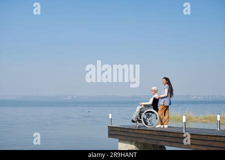 Foto di una nipote che si trova accanto a sua nonna in sedia a rotelle sul molo mentre entrambi si godono lo splendido panorama dell'ampio lago blu Foto Stock