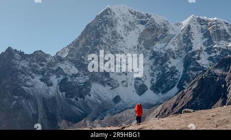 Una figura maschile solitaria che indossa uno zaino rosso si erge su un'aspra strada di montagna, con cime innevate sullo sfondo Foto Stock