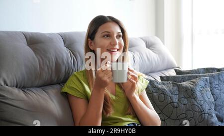 Bella donna in abiti informali seduta comoda sul divano che beve cioccolata calda ama il fine settimana, appartamento moderno, concetto di vacanza. ph. Orizzontale Foto Stock