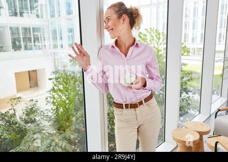 Donna d'affari felice che tiene una tazza di caffè appoggiata alla finestra Foto Stock