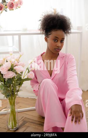 Giovane donna africana pensiva in pigiama rosa seduta sul pavimento vicino a un bouquet di fiori sullo sfondo della finestra. Foto Stock