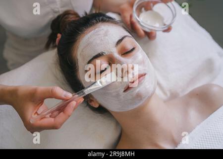 Vista dall'alto di una femmina anonima che applica la maschera facciale sul viso mentre si siede sul letto con gli occhi chiusi Foto Stock
