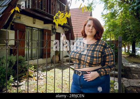 giovane donna latina di etnia argentina con capelli rossi e abiti casual in posa fuori dalla casa guardando la macchina fotografica, copiare Foto Stock