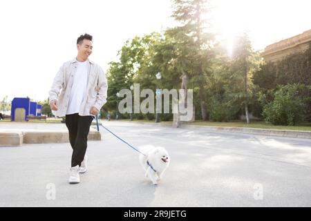 Felice giovane asiatico in abiti casual con adorabile cane bianco Pomeranian Spitz che cammina nel parco nelle giornate di sole Foto Stock