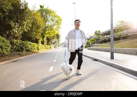 Allegro di un giovane asiatico in abiti informali con adorabile cane bianco Pomeranian Spitz che cammina per strada nelle giornate di sole Foto Stock