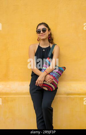 Giovane donna con outfit nero e occhiali da sole in piedi vicino a una parete gialla con colorata borsa a maglia e guardando lontano Foto Stock