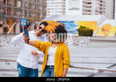 Giovane coppia polirazziale positiva ed elegante in abiti casual che scatta selfie sullo smartphone mentre si trova vicino alla ringhiera in città nelle giornate di sole Foto Stock