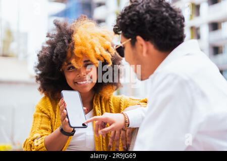 Felice femmina nera con i capelli gialli afro in abiti casual sorridente e guardando un amico anonimo maschio mentre condivide lo smartphone e si siede su sfocato Foto Stock