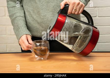 Primo piano di una donna che versa acqua calda da un bollitore in una tazza di vetro trasparente. una tazza di tè alle erbe appena fatto. Il processo di preparazione del tè o della cera di tè Foto Stock