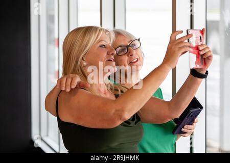Donna matura felice e madre in pensione in piedi vicino alla finestra e scattano un autoritratto sullo smartphone mentre si abbracciano Foto Stock