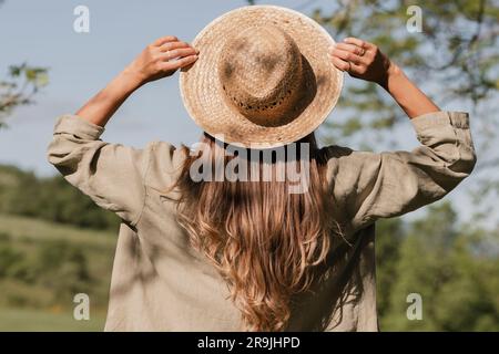 Vista posteriore di una donna irriconoscibile con cappello di paglia e abbigliamento casual rilassandosi sul campo verde e ammirando la vista panoramica del paesaggio Foto Stock
