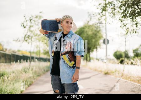 Giovane donna timida e positiva in un abbigliamento casual da maschiaccio con lo skateboard posizionato sulle spalle della strada e guardando lontano Foto Stock