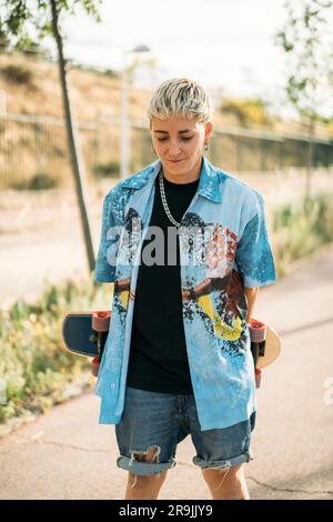 Giovane donna timida e positiva in un abbigliamento casual da maschiaccio che tiene lo skateboard con le mani dietro la schiena mentre si trova in strada e guarda verso il basso Foto Stock