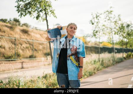 Giovane donna timida e positiva in un outfit casual da maschiaccio con lo skateboard posizionato a spalla sulla strada guardando lontano Foto Stock