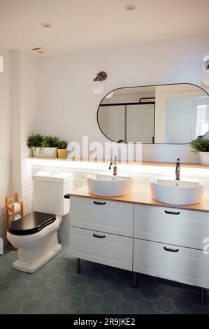 Design interno del bagno con lavandini in ceramica bianca rubinetti su armadietto in legno vicino alla ciotola igienica collocata in un angolo del pavimento piastrellato di granito Foto Stock