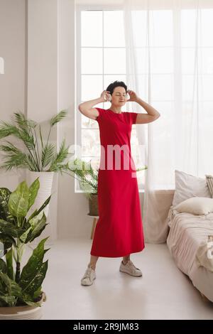 Tutta la lunghezza di una donna positiva di mezza età in abito rosso che balla con le braccia alzate mentre ti godi la musica in una camera da letto chiara con piante verdi Foto Stock