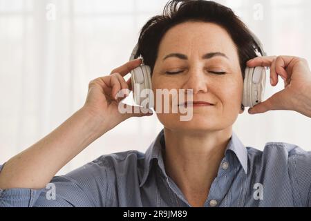 Vista frontale di una donna di mezza età con i capelli scuri corti in camicia blu che ascolta la musica con le cuffie wireless Foto Stock