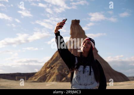 Ritratto di una giovane viaggiatrice allegra in abiti casual che scatta selfie sullo smartphone mentre si stende sullo sfondo delle montagne e del cielo blu a Nav Foto Stock