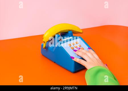 Alto angolo di donna anonima con mano su un telefono in plastica vecchio stile con banana su superficie arancione su sfondo rosa Foto Stock