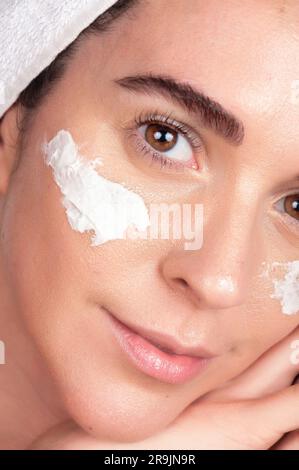 Primo piano del raccolto sorridente giovane femmina con un asciugamano avvolto sui capelli appoggiandosi la testa a mano e guardando la macchina fotografica con crema per maschera applicata Foto Stock