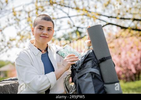 Ritratto di una giovane escursionista tatuata e dai capelli corti in abiti informali prendendo una mappa dallo zaino con tappetino fitness e guardando lontano con la natura al b Foto Stock