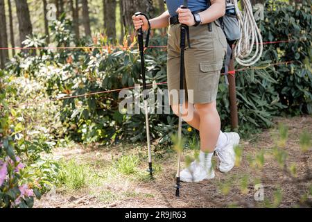 Vista ravvicinata della giovane donna con tracker per il fitness e zaino che tiene i bastoni da trekking mentre cammina lungo il sentiero nella foresta verde sfocata, ricollegandosi con Foto Stock