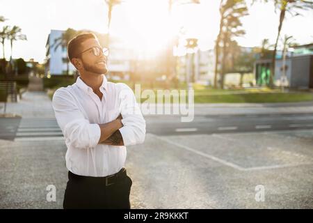 Felice uomo d'affari europeo da sogno, vestito in modo formale, in piedi all'aperto con le braccia piegate e guardando lontano lo spazio vuoto Foto Stock