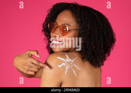 Felice giovane donna riccia nera con occhiali da sole che punta il dito contro la crema solare alle spalle Foto Stock