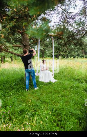 Un videografo professionista per matrimoni realizza un film con una sposa su un'altalena di corda in una pineta Foto Stock