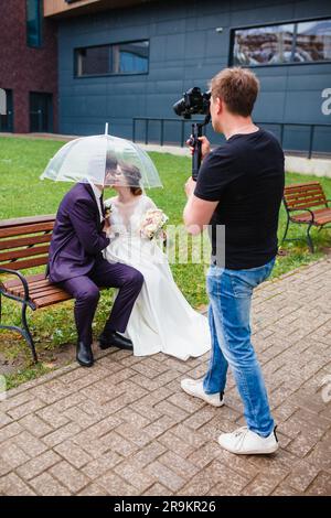 Un videografo professionista per matrimoni registra un film con gli sposi sposi su una panchina sotto un ombrello Foto Stock