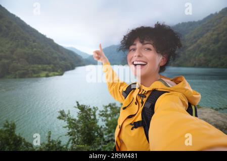 Bella donna che scatta selfie e mostra i pollici vicino al fiume in montagna Foto Stock