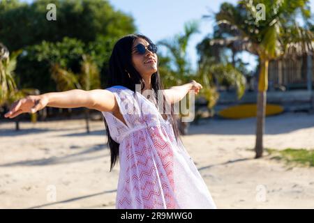 Felice donna birazziale con occhiali da sole e abito da spiaggia bianco che balla sulla spiaggia soleggiata sorridendo Foto Stock