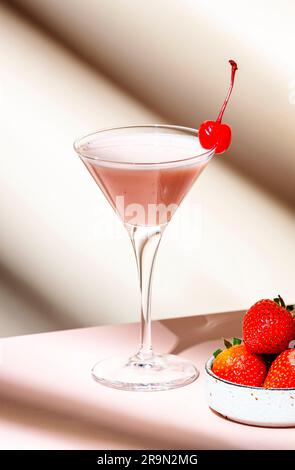 Cocktail estivo di grandi martini con vodka, liquore, panna, fragole e ghiaccio in bicchiere con ciliegia rossa da cocktail. Sfondo rosa beige Foto Stock