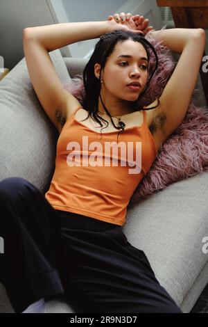 ritratto di una giovane donna con le braccia alzate e i capelli delle ascelle, corpo naturale, rilassante sul divano Foto Stock
