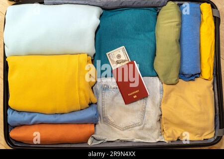 Vista dall'alto verso il basso di una valigia con abiti da donna ben riposti, un passaporto e una pila di dollari. Preparazione per il viaggio, le vacanze estive. Trave d'aria Foto Stock