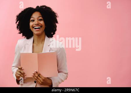 Ritratto di una giovane donna di colore che tiene un notebook, sorride e sorride, sullo sfondo rosa dello studio Foto Stock