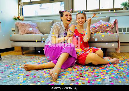 Due amici che fanno festa a casa seduti sul pavimento con dei soffiatori da festa con dei coriandoli che cadono intorno a loro. LGBT, non binario Foto Stock