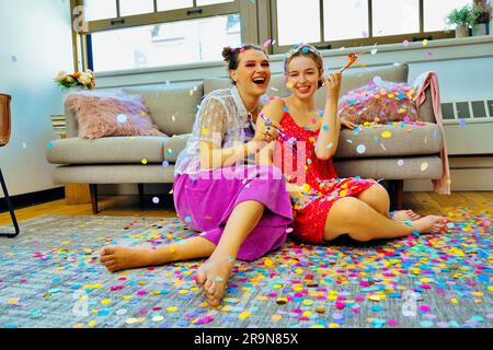 Due amici che fanno festa a casa seduti sul pavimento con dei soffiatori da festa con dei coriandoli che cadono intorno a loro. LGBT, non binario Foto Stock