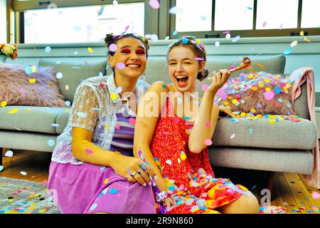 Due amici che fanno festa a casa seduti sul pavimento con dei soffiatori da festa con dei coriandoli che cadono intorno a loro Foto Stock