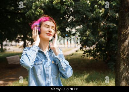 Una ragazza con i capelli rosa e le cuffie ascolta la musica nel parco in estate Foto Stock