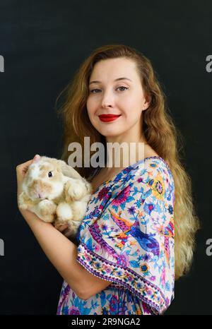 Bella donna russa con lunghi capelli biondi che tiene in mano un animale domestico di coniglio contro il muro nero Foto Stock