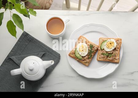 Deliziosi sandwich con tonno, verdure, uovo sodo e tazza di caffè su un tavolo in marmo bianco al chiuso, piatti. Colazione gustosa Foto Stock
