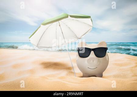 Sconti per le vacanze. Salvadanaio con occhiali da sole e ombrellone sulla spiaggia sabbiosa vicino al mare Foto Stock