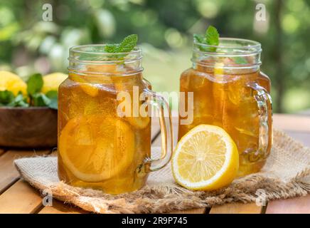 Tè ghiacciato in vasetti di vetro con fettine di limone e menta all'aperto su un tavolo di legno con sfondo naturale Foto Stock