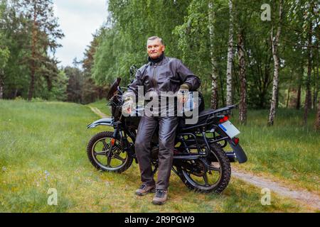 Motociclista anziano che si addossa a una moto nella foresta estiva. Uomo in pelle che si rilassa godendosi la natura dopo il giro Foto Stock