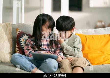 piccolo fratello e sorella di bambini asiatici seduti sul divano della famiglia a casa utilizzando un tablet digitale insieme Foto Stock