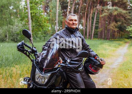 Biker anziano appoggiato su motobike nella foresta estiva. Uomo in pelle vestiti in piedi con le braccia incrociate godendo la natura. dopo il giro Foto Stock