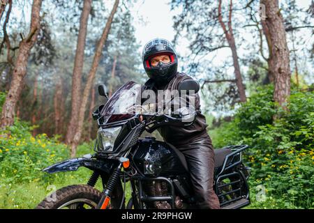 Motociclista senior in moto all'aperto. Uomo in pensione con giacca in pelle che riposa dopo aver guidato nella foresta estiva appoggiandosi al manubrio. Viaggio estivo in auto Foto Stock